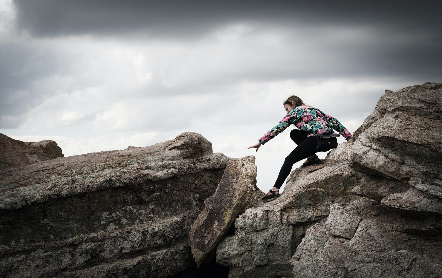 An individual in a floral sweater perched mid-climb on some rocks while reaching towards another section of rock before moving forward. Cloudy skies behind.