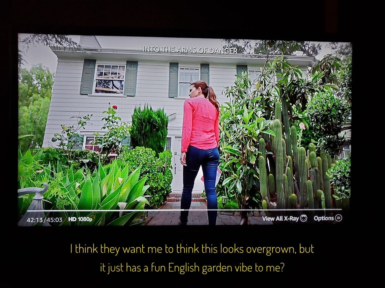 Laura approaching the house through the front yard, which has a big cool cactus, a rhododendron, a rosebush, and a bunch of other plants, captioned "I think they want me to think this looks overgrown, but it just has a fun English garden vibe to me?"