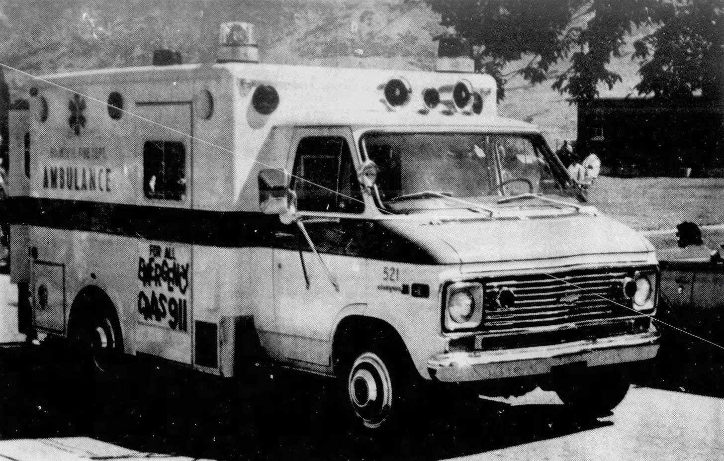 A black-and-white newspaper photograph of a new ambulance, parked in front of an expanse of lawn on a bright day with hills in the background.