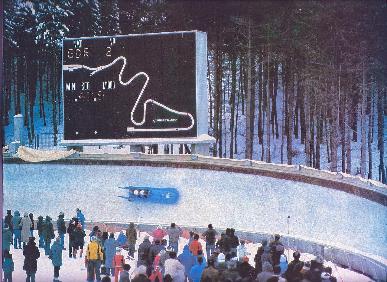 A photo from the Sarajevo Winter Olympics showing a blue two man bobsleigh hurtling down the run while a crowd looks on. 