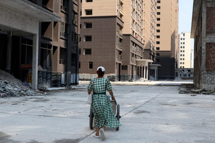 A woman in a green checked dress and headscarf pushes a cart down a concredte road between unfinished buildings