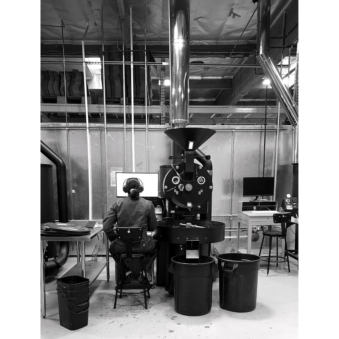 A black and white photo of a commercial coffee roasting machine with a drum and hopper. To the left a man in a jumpsuit wearing oversized headphones and his hair back in a ponytail stairs a computer screen monitoring the coffee bean roasters temperature.
