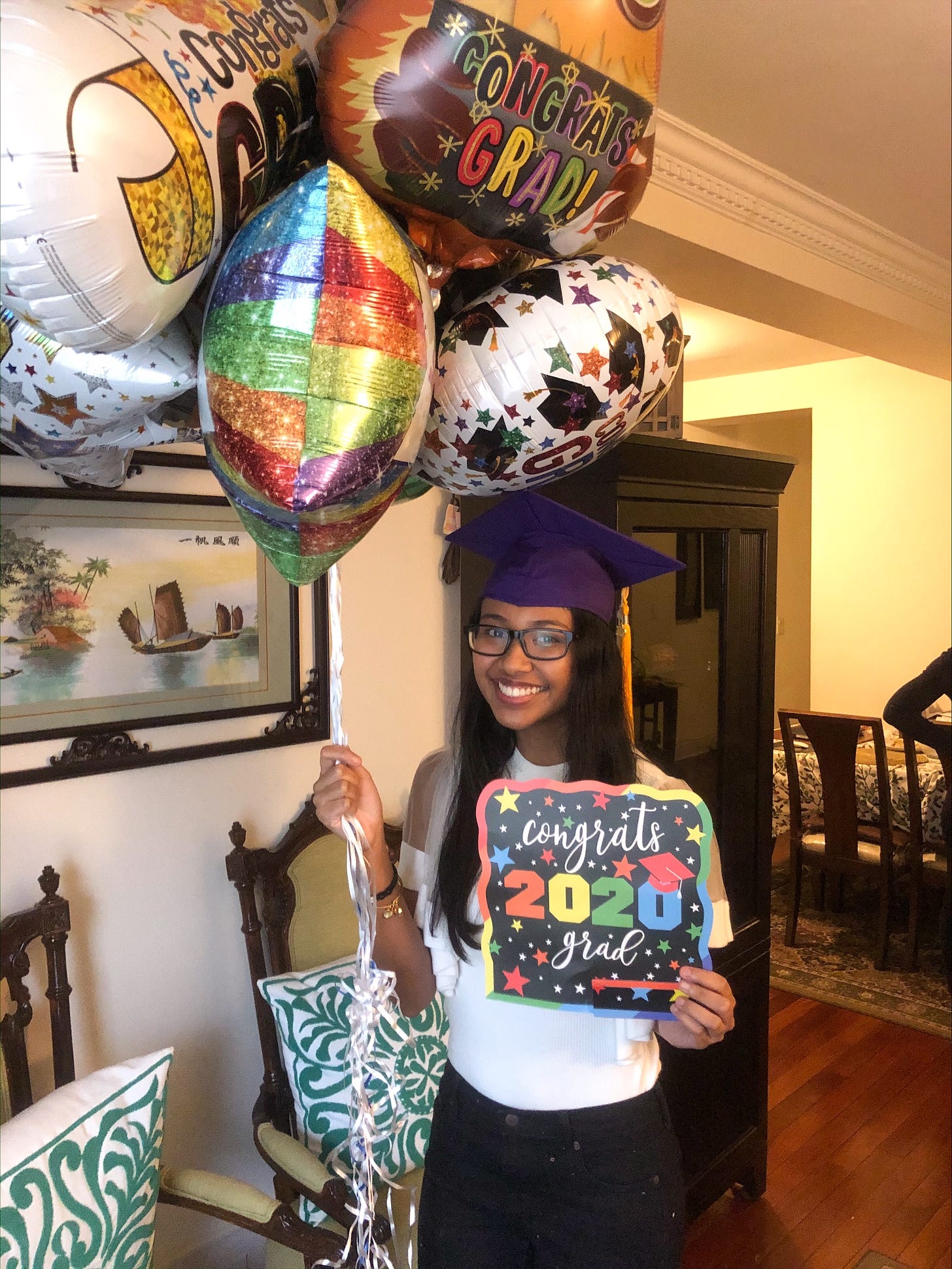 Melissa, standing in her living room with a purple grad cap on. She's holding grad balloons and a sign that says, "Congrats 2020 grad."