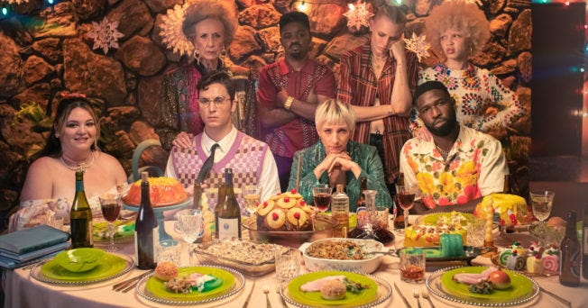 A diverse group of eight people pose at a holiday dinner table, looking uncomfortable like they are relatives who don't enjoy each other's company