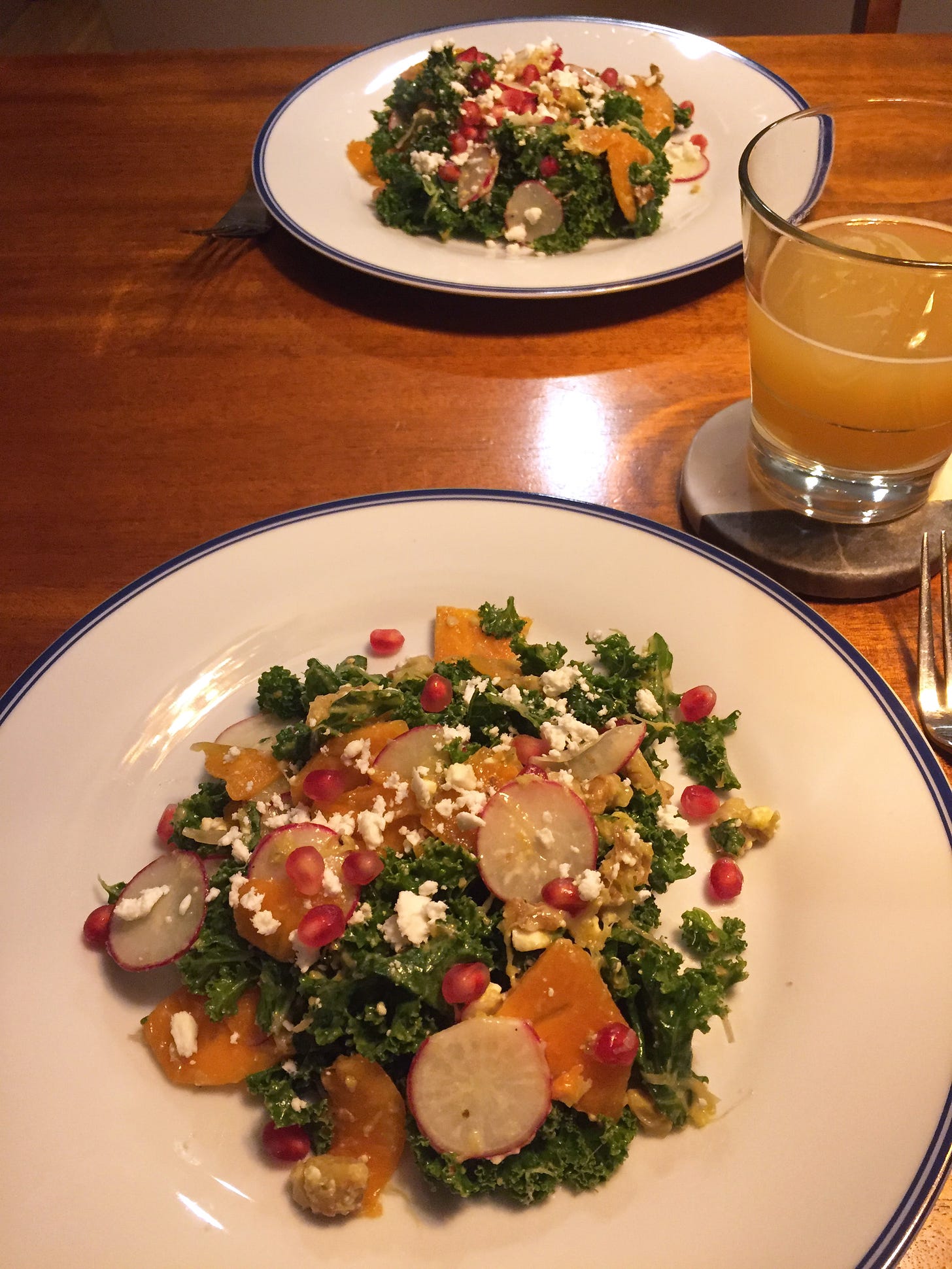 Across from each other are two white plates of kale salad, with orange slices of persimmon and thin radish rounds throughout. Feta is crumbled overtop, and pomegranate seeds are scattered over it.
