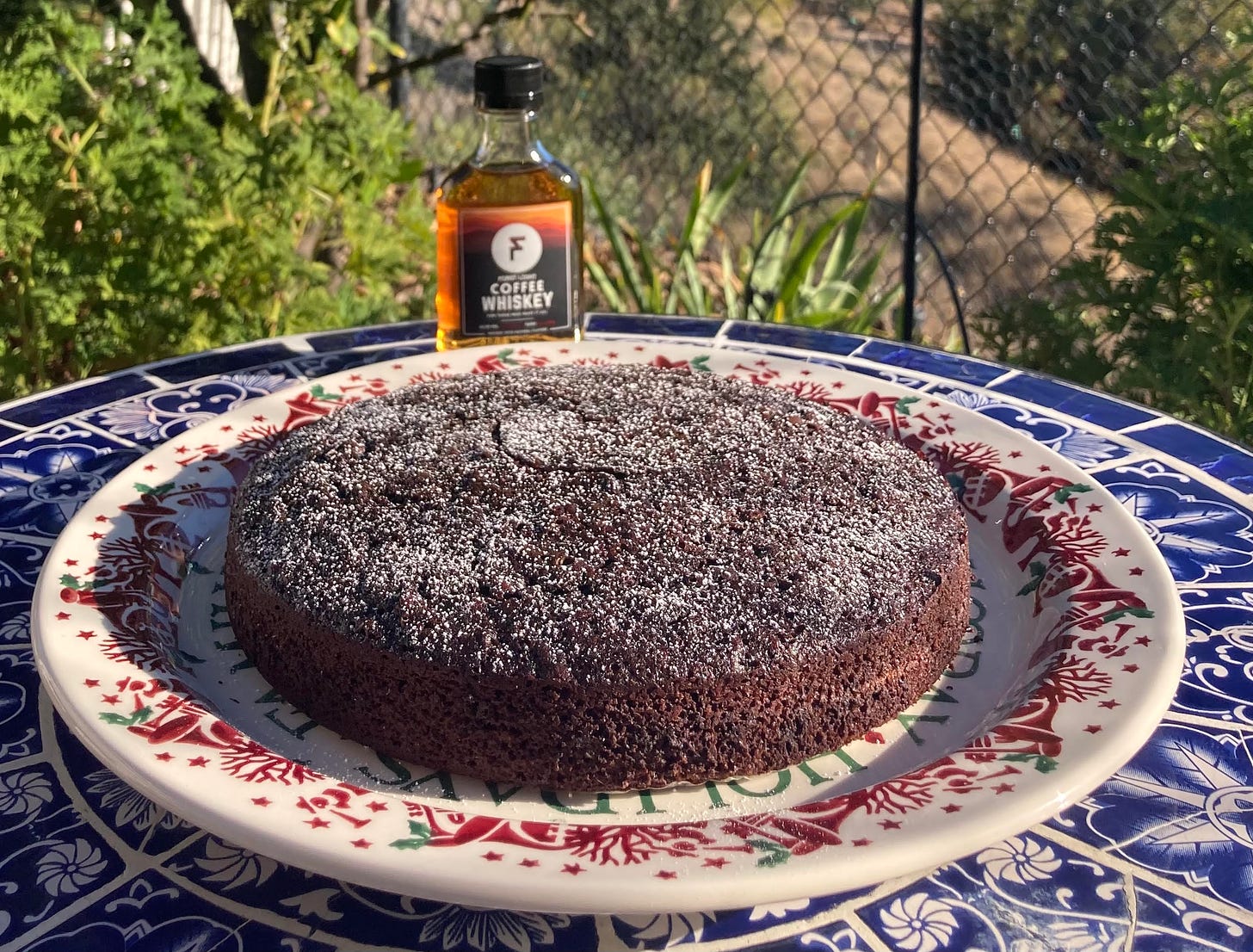 A flat, round chocolate cake dusted with confectioner's sugar on a white plate with a ring of red Christmas decorations around the edge. A bottle of First Light Coffee Whiskey is on the table just beyond the cake. The table is blue with a white floral pattern. Outside. Close-up photo.