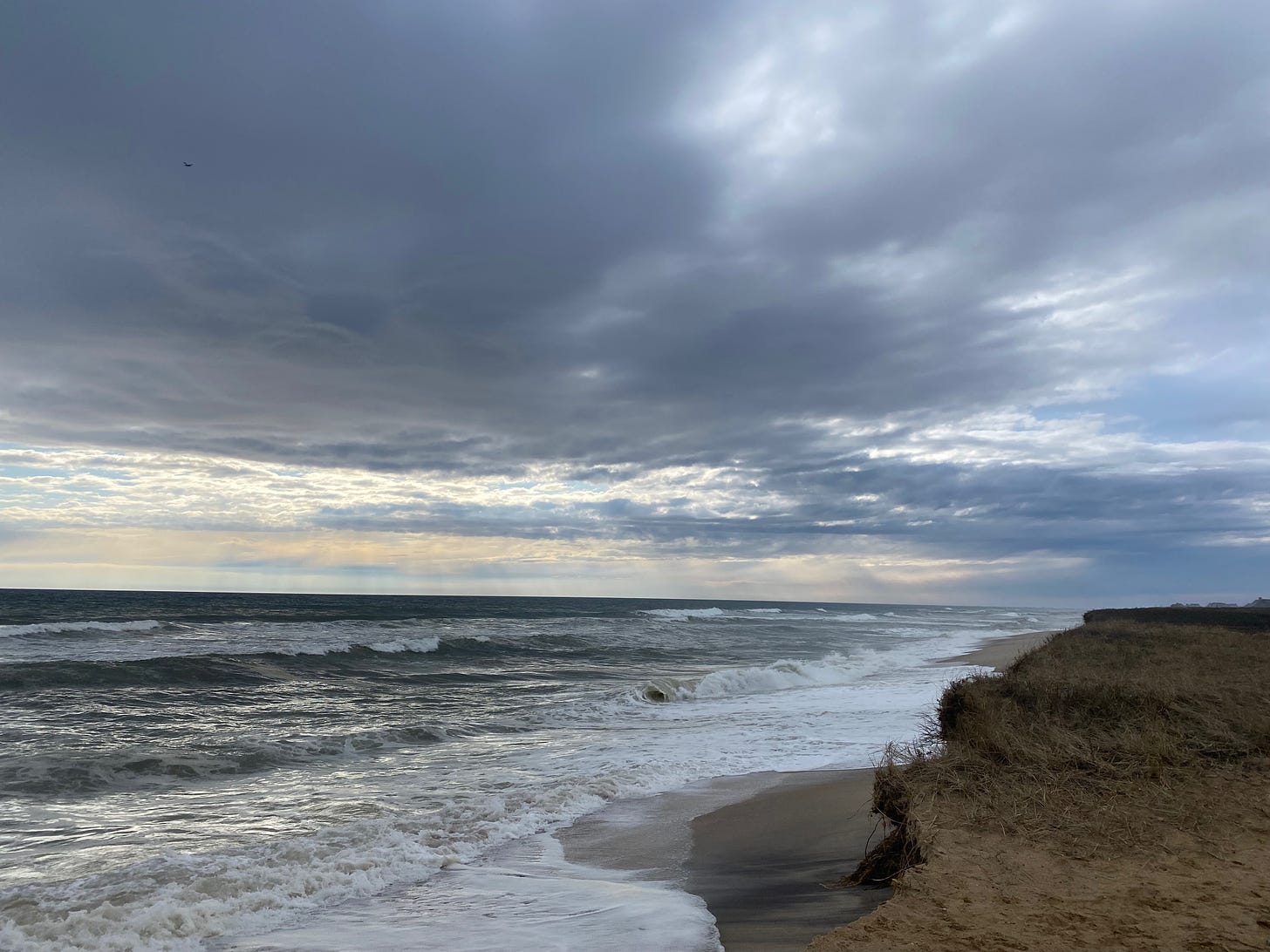 A view of a stormy beach. The waves are breaking white and foamy on the shore, almost reaching the windswept dunes; the water is dark. The sky is full of purple and silver clouds; the sun is behind them, but several rays shine down on the horizon. 