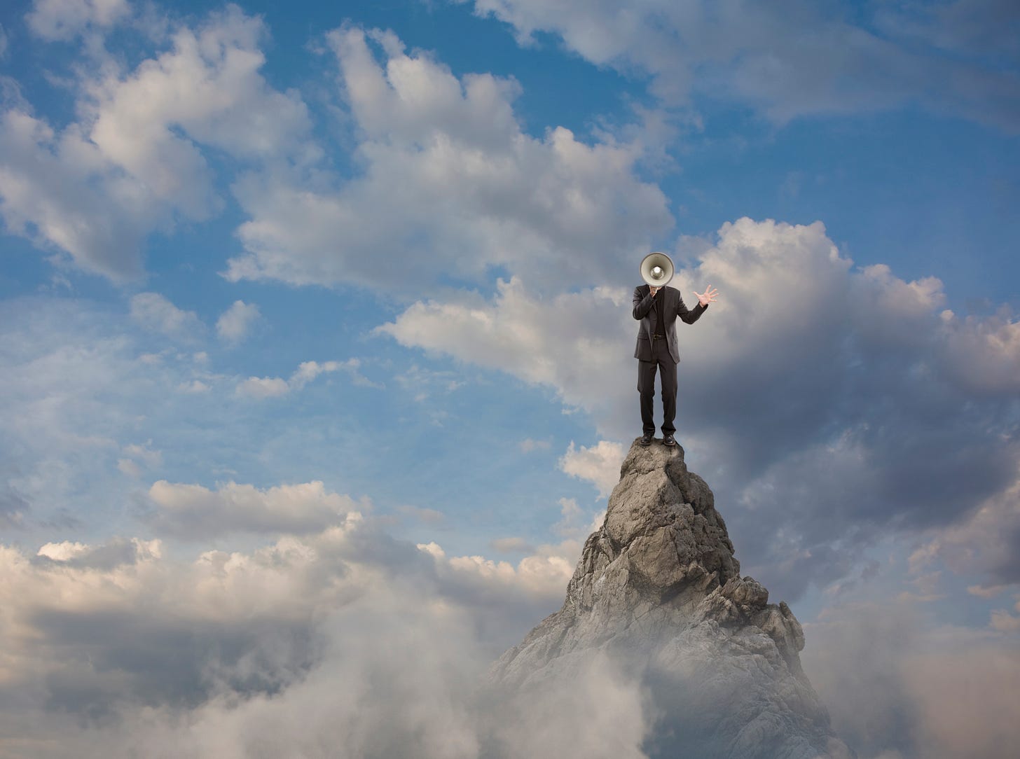 A person in a suit standing on a rocky  mountain pinnacle speaking into a hand held megaphone
