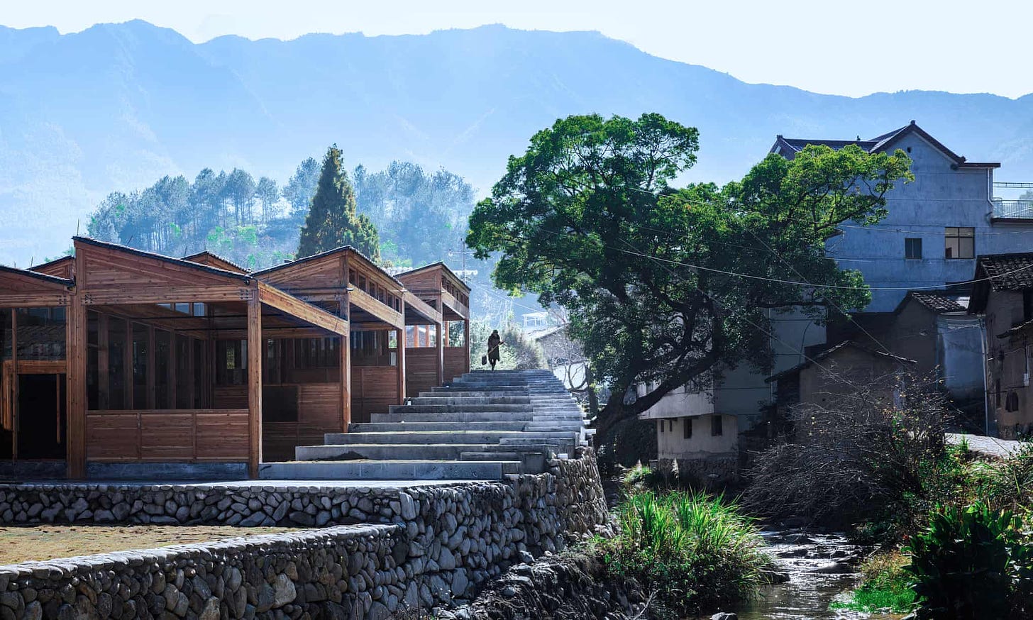 A colour photo of a Chinese mountain village. A series of wooden pavilions step down a slope next to a stream, with mountains and forest in the background