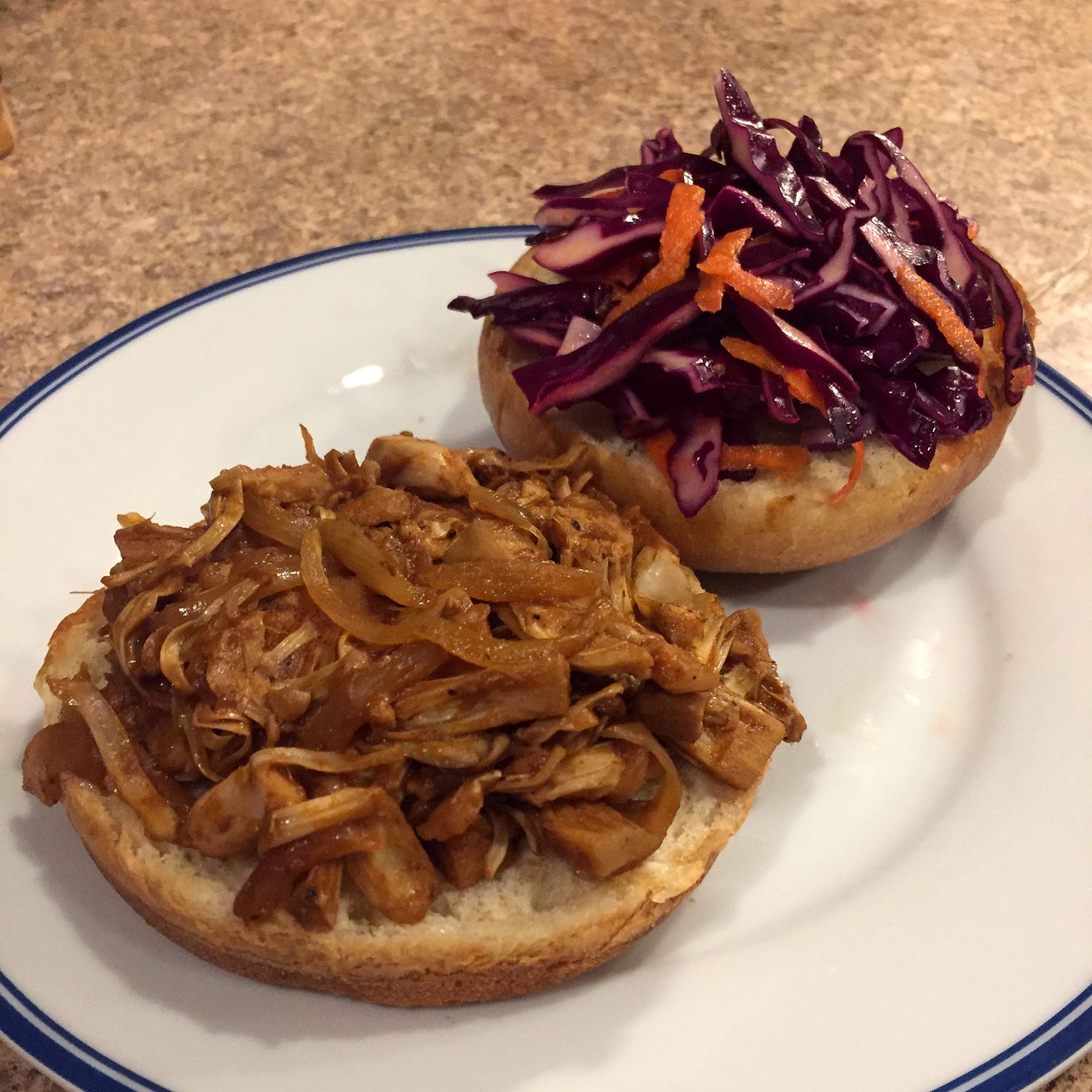 two halves of a brioche bun on a white plate. The bottom has pulled jackfruit and onions in barbecue sauce, and the top has red cabbage and carrot slaw.