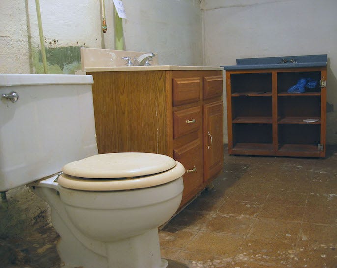 A dirty large toilet on a bare floor of a cinderblock walled room with a standalone vanity and shelf unit. There is a sign tacked above the faucet that says "Not connected"