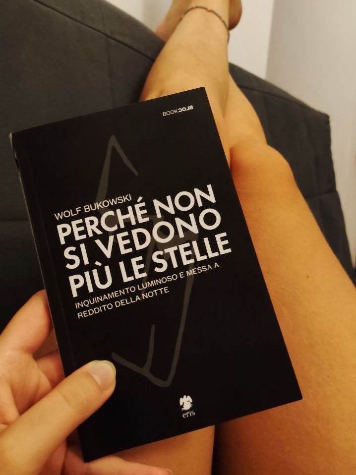 Una mano tiene un libro. Si intravedono le gambe di una persona, distesa sul divano. I piedi sono appoggiati in alto, sullo schienale del divano. Il libro, con la copertina nera, è "Perché non si vedono più le stelle. Inquinamento luminoso e messa a redditto della notte" di Wolf Bukowski.