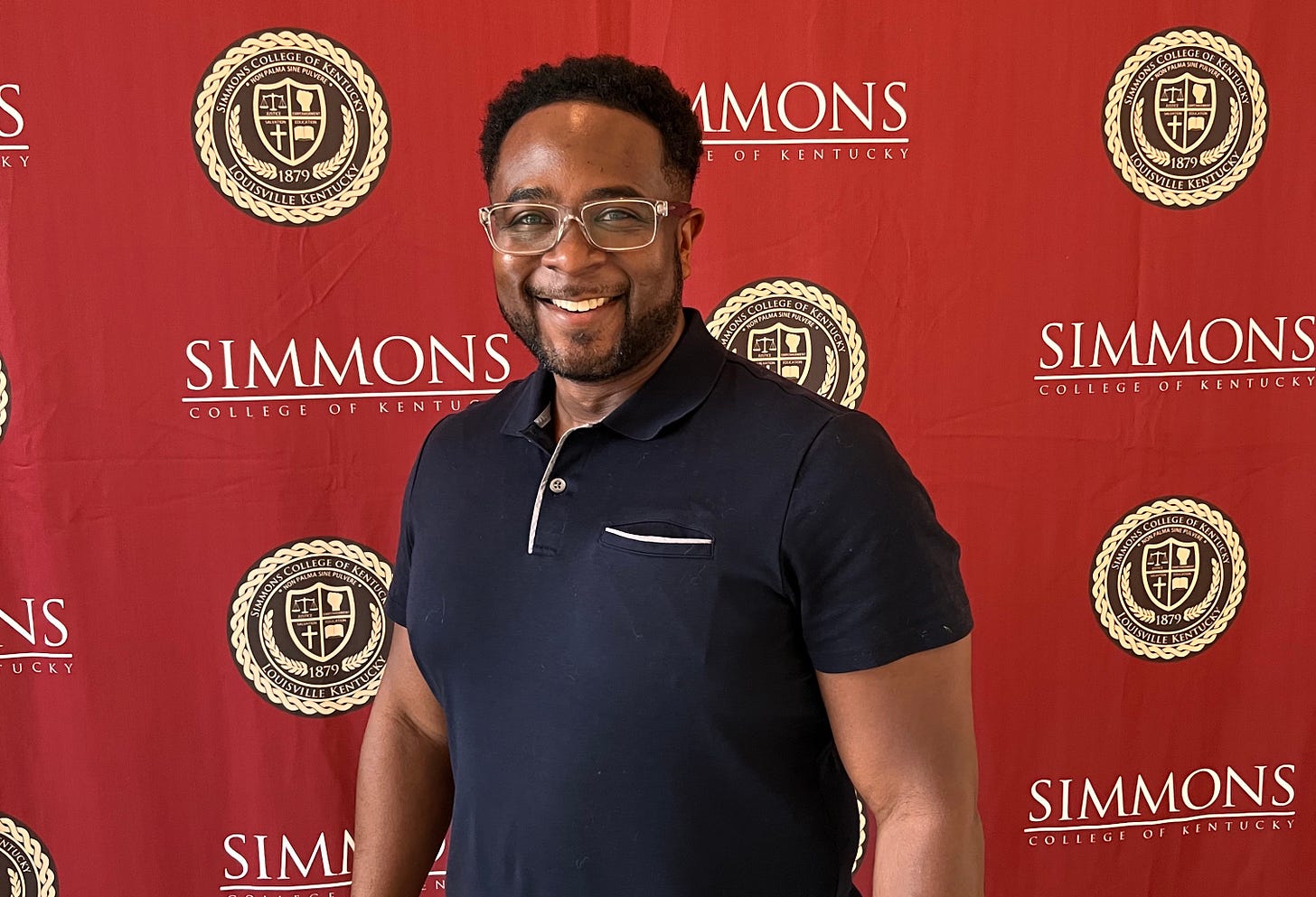 Jemar Tisby smiling and standing in front of a backdrop that says Simmons College