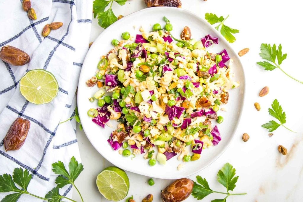white rimmed plate with colorful chopped salad, on the side is a dish towel with fresh lime, dates, fresh parsley and some additional pistachios and herbs strewn about on the white countertop. 