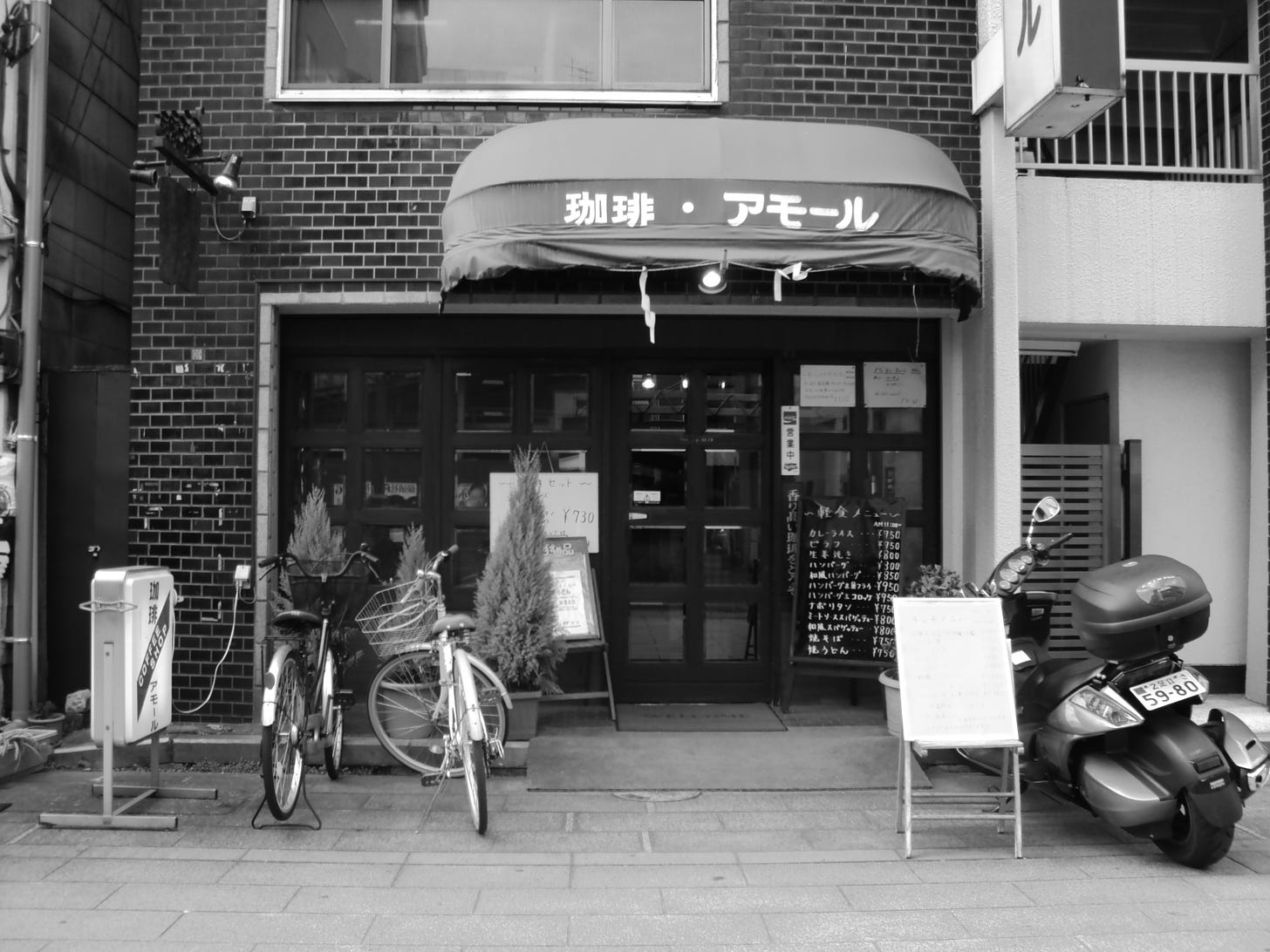 View of a shop from the outside. Two bicycles and a moped are parked right in front of it. Image is in black and white.
