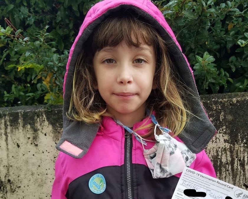7-year-old kid in a pink hoodie with a mask around her neck holds her new Covid-19 vaccine card and wears a vaccinated sticker