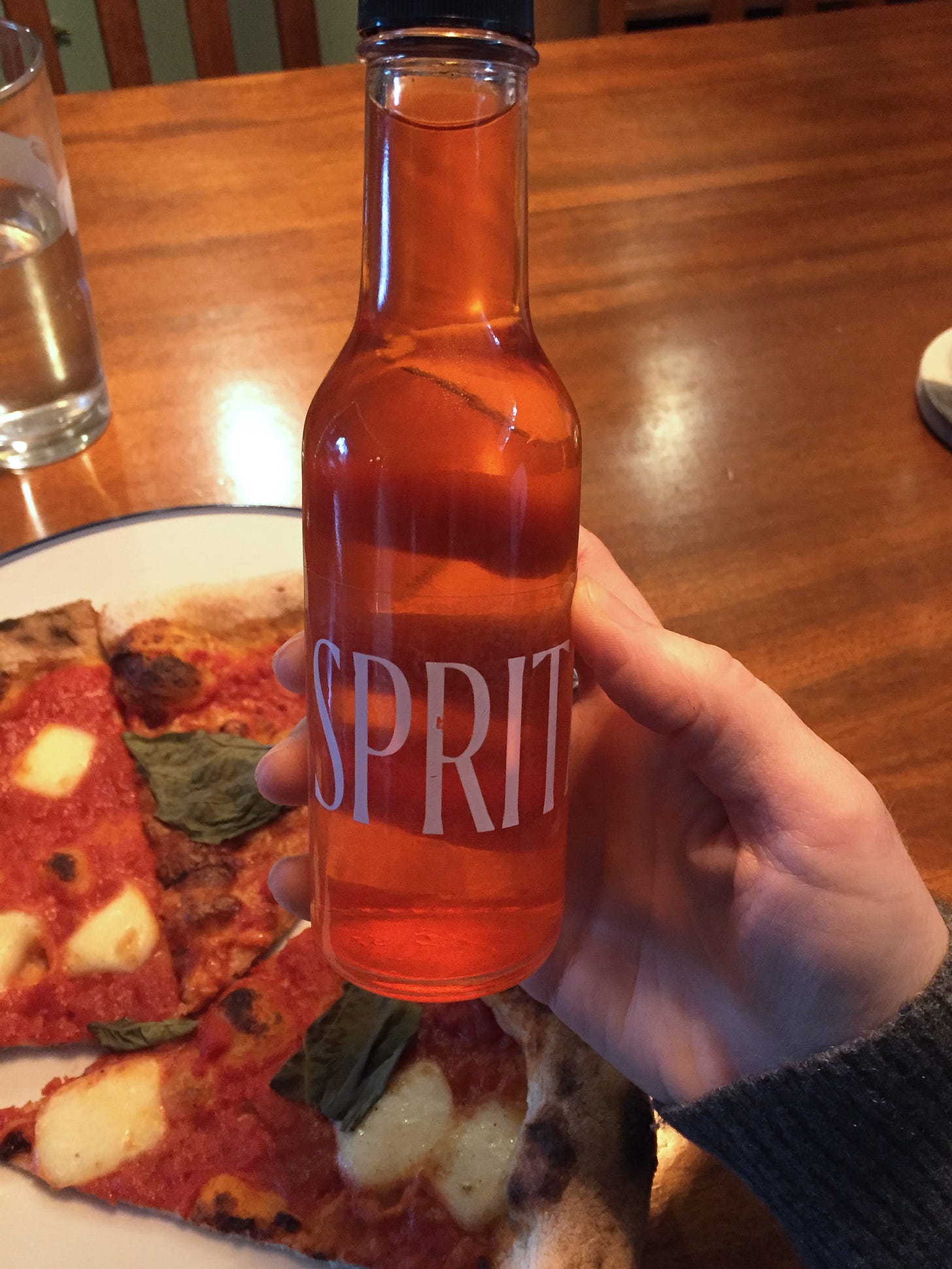 a tiny bottle of reddish Aperol spritz being held in front of a plate of margherita pizza