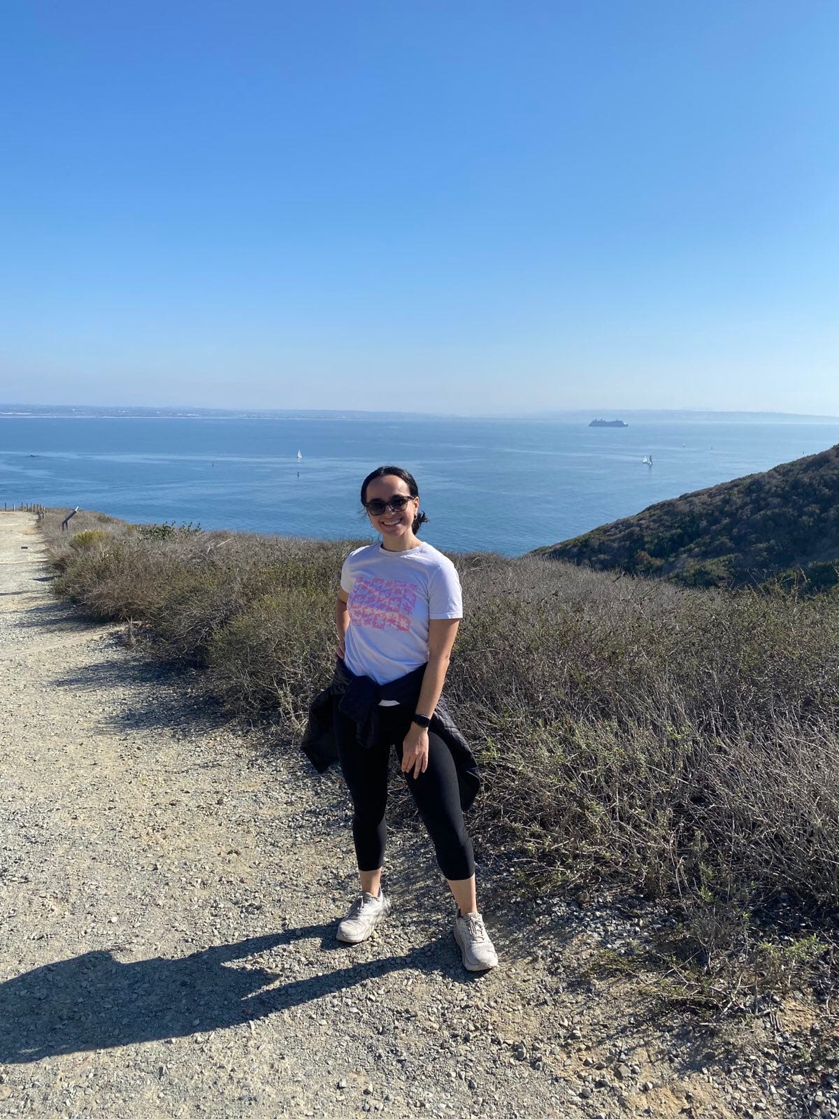 Alana stands in the middle of the photo, smiling and in workout gear. She's standing on a gravel and dirt path with brush next to her. Behind her is a full view of the sky and ocean.