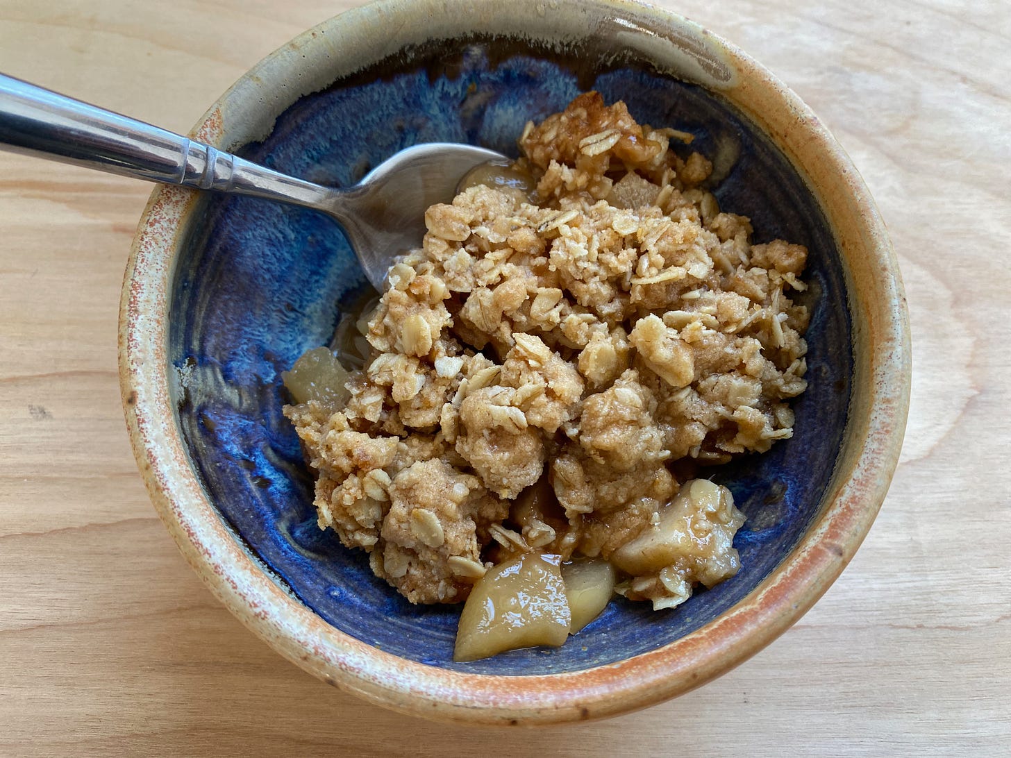 A small blue ceramic bowl of apple crisp with a spoon resting in it.