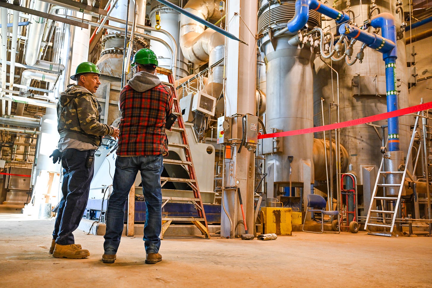 two men in green hard hats stand looking at a maze of pipes, elbows, and connections inside a wastewater treatment plant. the photographer stands behind them and captures them in mid conversation, the man on the left pointing to the equipment as he talks to the man on the right.
