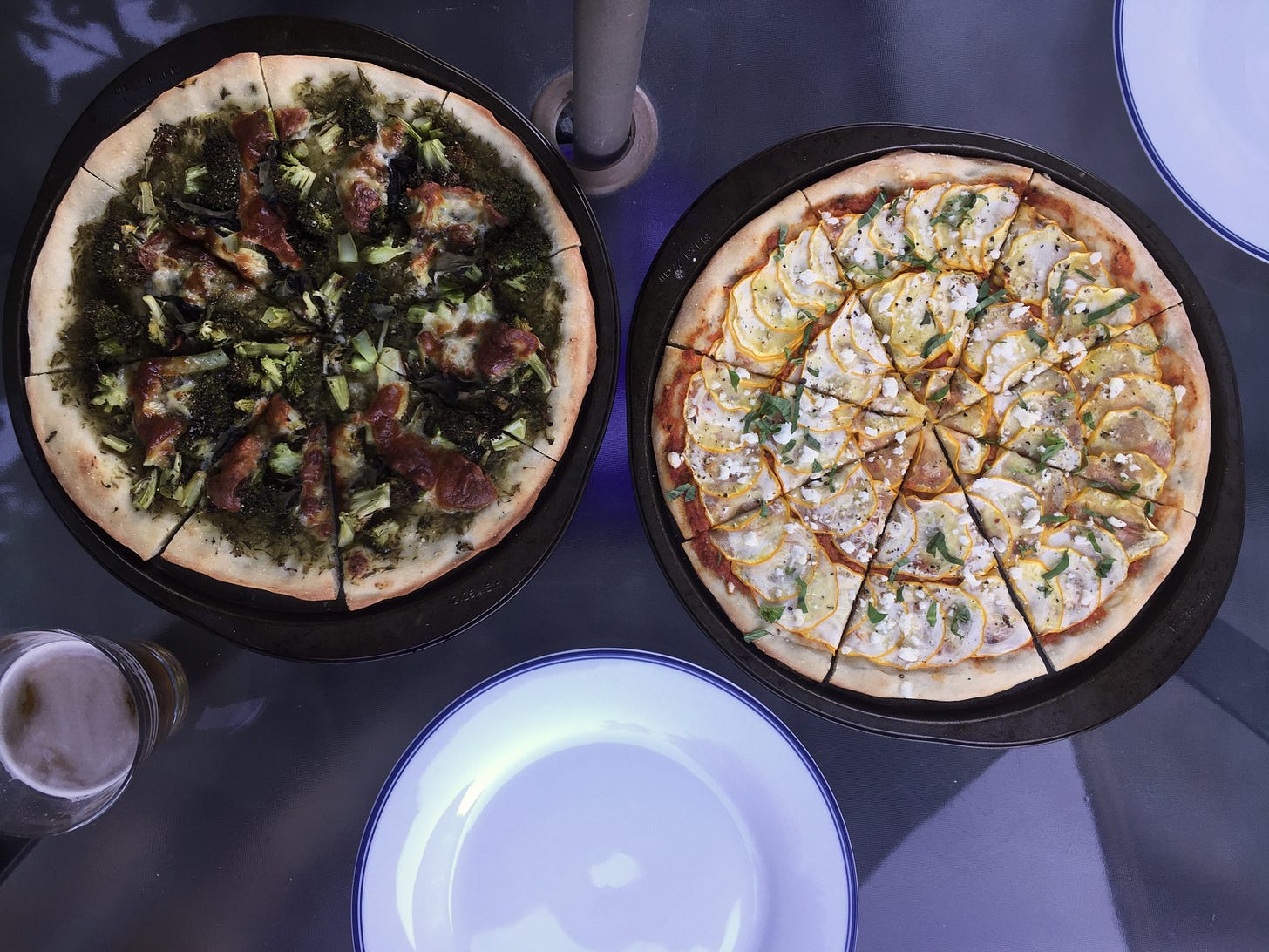 two pizzas in their pans. On the left, a green garlic scape pesto with pieces of broccoli and browned patches of provolone. on the right, a red romesco base with concentric circles of overlapping yellow zucchini, and a dusting of feta and basil.