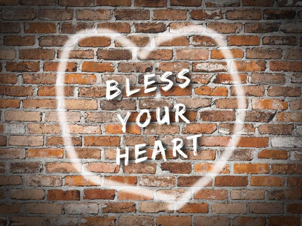 White graffiti spray painted on brick wall says "Bless Your Heart" inside a heart.