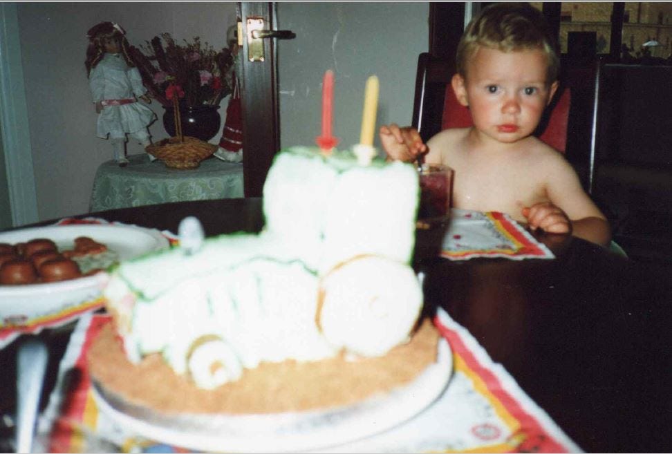 A two-year boy, with blonde hair, topless, sitting at a dark wood table. In front of him there's a birthday cake in the shape of a tractor.