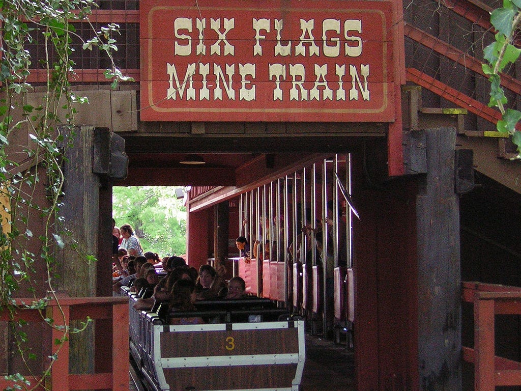 People preparing to ride a roller coaster. The sign above says, "SIX FLAGS MINE TRAIN".