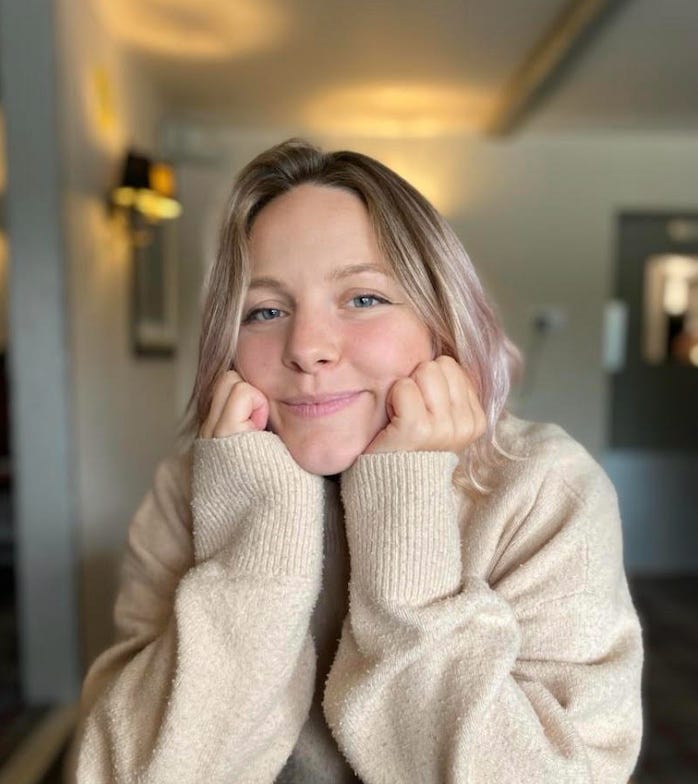 Ettie is smiling at the camera, with her head supported by her hands. She's a White woman, with blonde hair. She's wearing a cosy beige jumper.