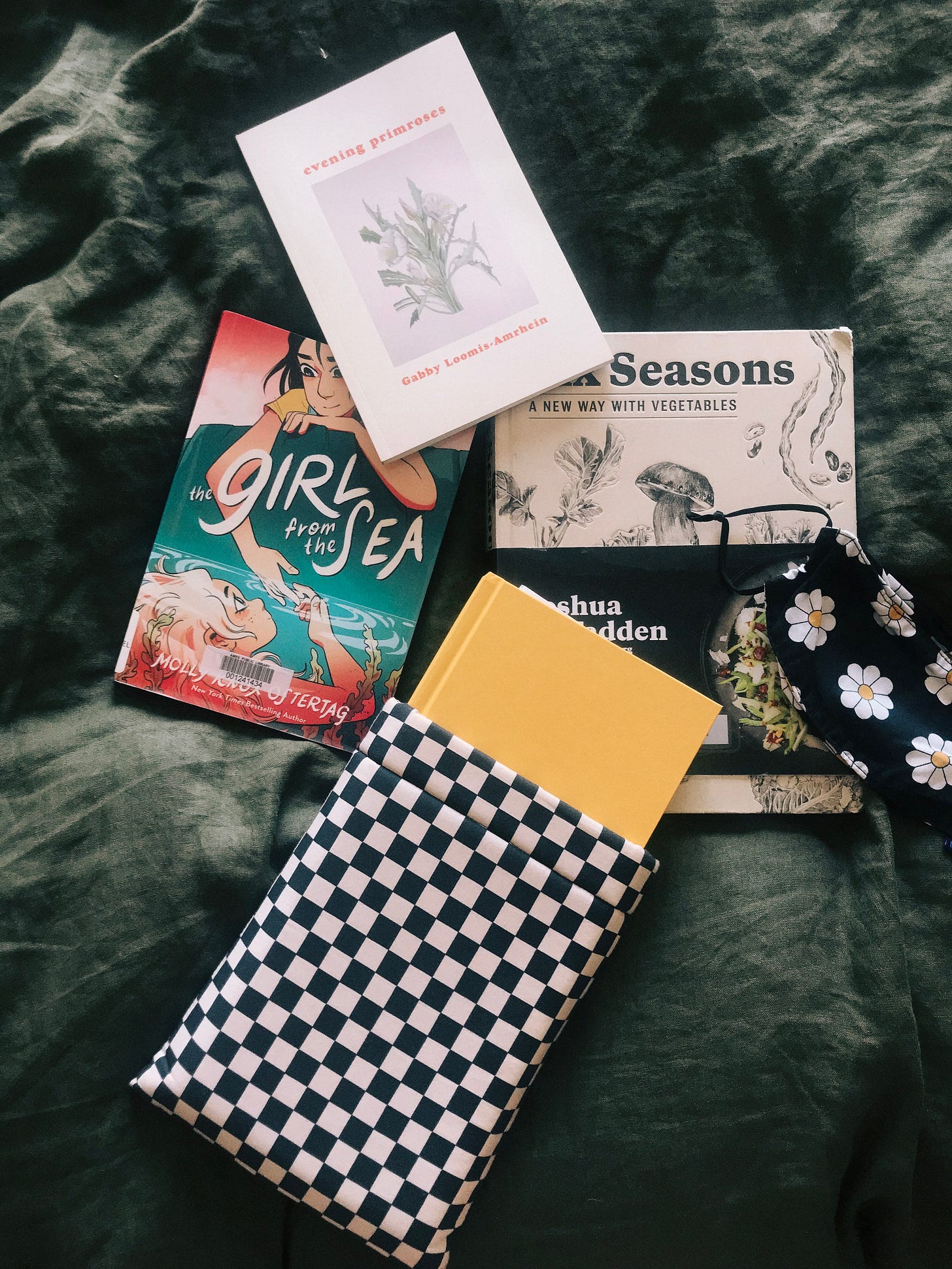 Four books on top of a dark green duvet. evening primroses is at the top of the diamond. On the left side is The Girl from the Sea. On the right side is Six Seasons. On the bottom is a yellow book with no title, sticking out from a black and white book sleeve. A black face mask with white flowers is also in the frame.