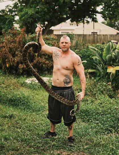 A photo of the front of muscular, tattooed, and mohawked Ryan Ausburn holding a snake up on display in an overgrown garden.