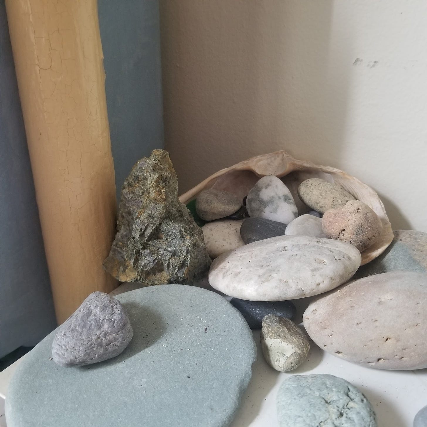 Image description: against a corner of an apartment by a narrow hot water pipe, someone has arranged a small and medium-sized rocks, some overlapping, some in a large clam shell.