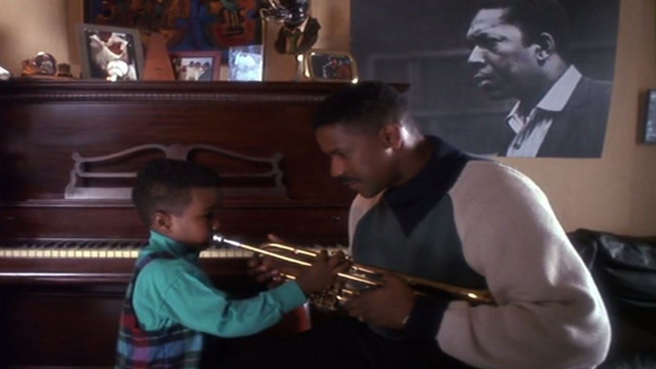 This image shows Bleek Gilliam (played by Denzel Washington) training his son, Miles (played by Zakee Howze) to master the trumpet near the end of Spike Lee's film Mo' Better Blues. Bleek and Miles stand before a piano and a large photograph of jazz legend John Coltrane. This image shows Bleek Gilliam (played by Denzel Washington) training his son, Miles (played by Zakee Howze) to master the trumpet near the end of Spike Lee's film Mo' Better Blues. Bleek and Miles stand before a piano and a large photograph of jazz legend John Coltrane.