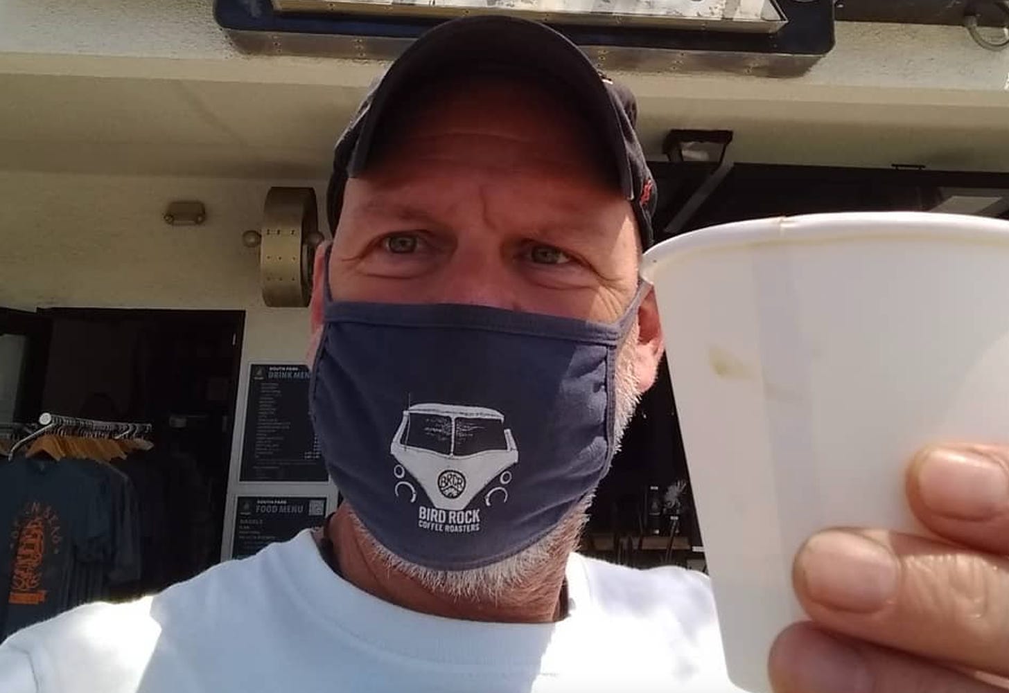 Selfie of a man in a baseball cap, wearing a blue face mask and holding a white takeout coffee cup up to the camera.