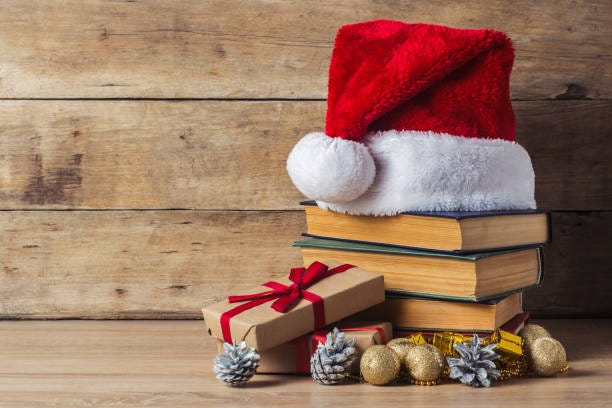 A planked background and laminate floor upon which lies a pile of vintage books. On top is a Santa hat made of red fur with a white pompom and white vedging. There's a brown paper parcel tied with red string propped against the pile of books. At their feet are pinecones sprayed silver, golden baubles and beads. This image encapsulates the theme of my newsletter which focuses on cookbooks with a seasonal theme