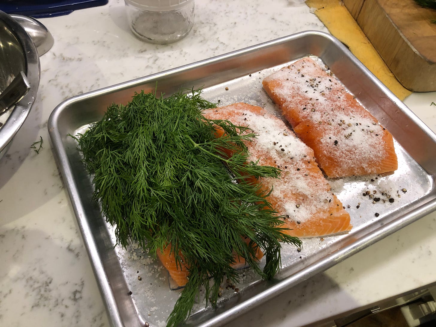 The salmon pieces, covered in the cure. One piece also has a large pile of dill on it, covering it completely.