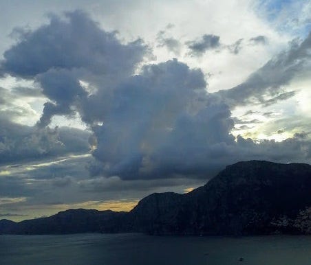 Dusk over the Amalfi Coast. The darkening sky is full of puffy clouds and below, the mountains are dark silhouettes.