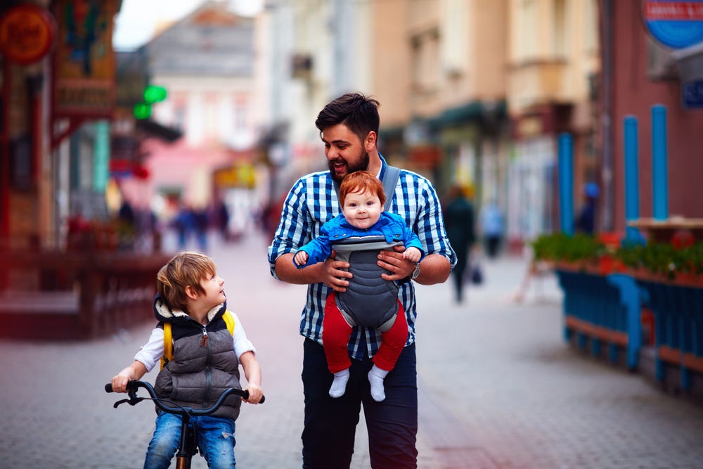 Father walking down the street with two young sons. One is on a bike, father and son making eye contact. The other child is in a baby carrier strapped to his chest. The street is cobblestone and lined with cafes, soft focus.