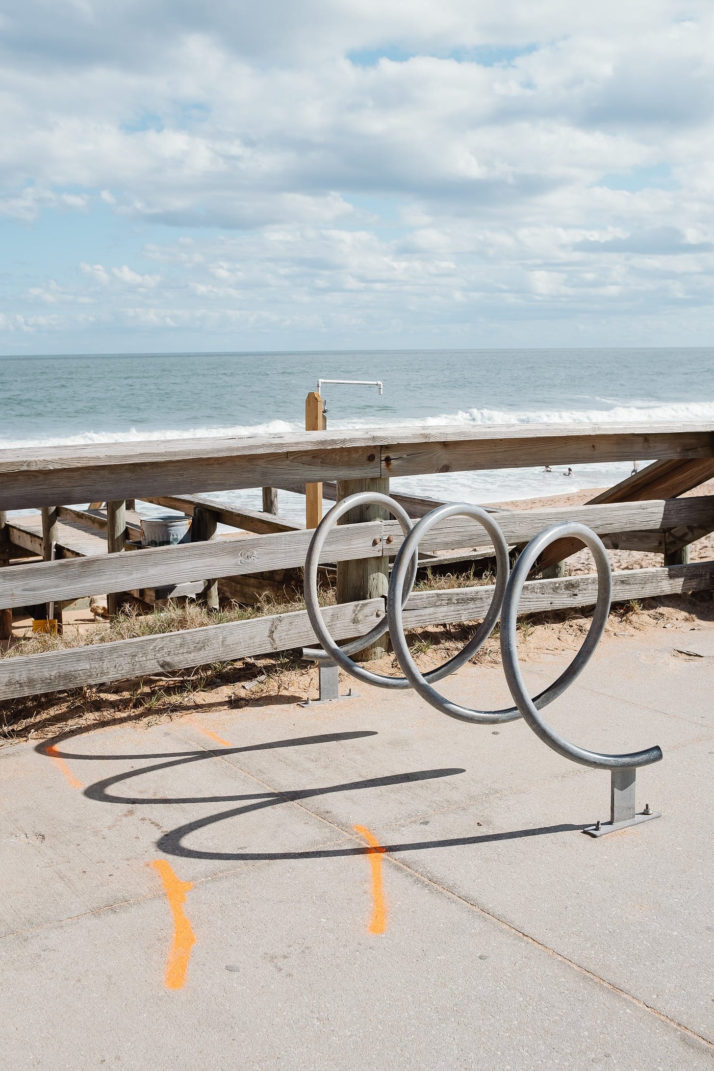 A fence with a beach and the sea behind, a curly bike stand in the foreground with a curly shadow on the floor.
