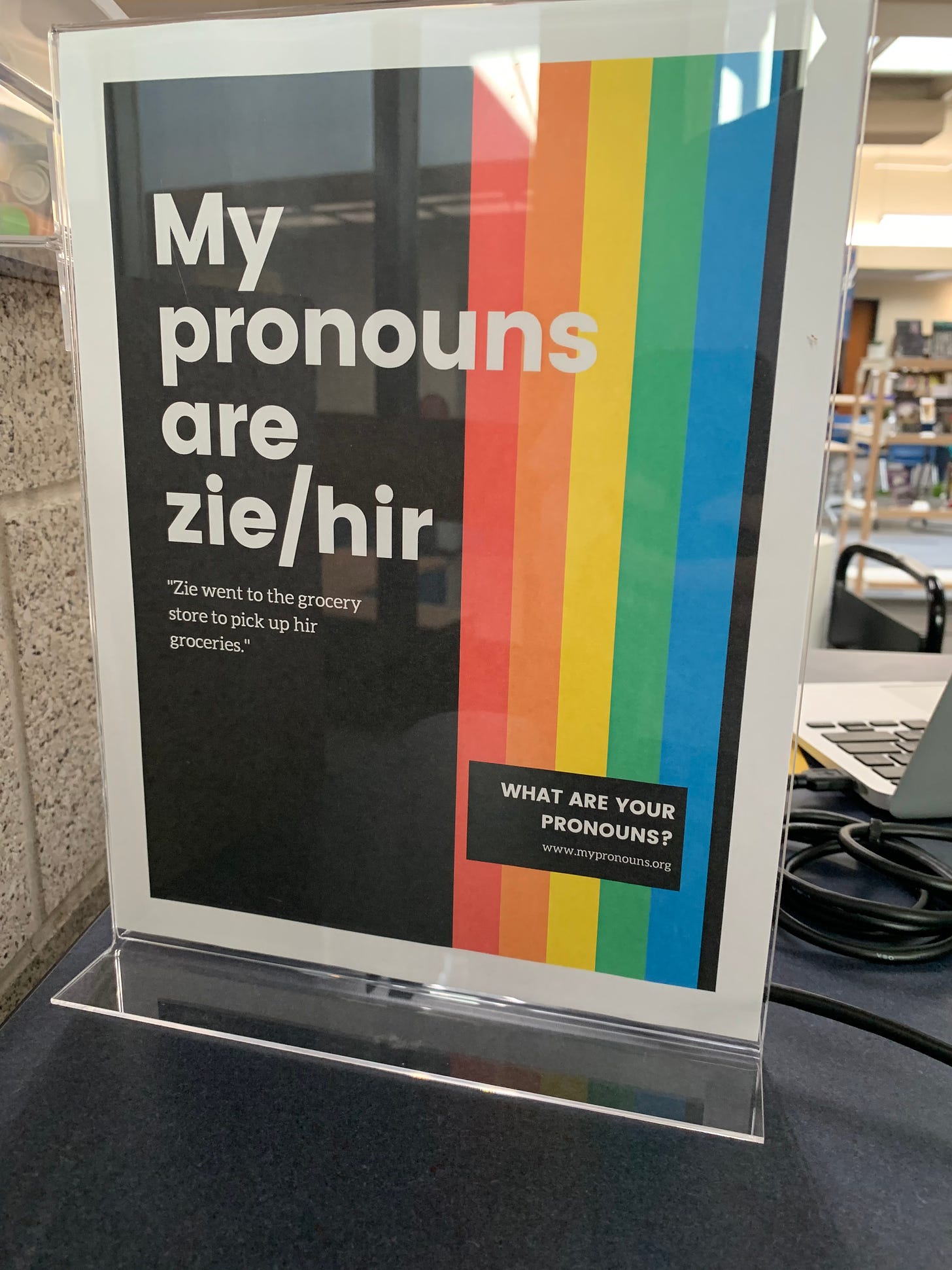 A poster in a plastic cover has a rainbow over black. The white text reads “My pronouns are zie/hir. “Zie went to the grocery store to pick up hir groceries.” What are you pronouns? www.mypronouns.org” 