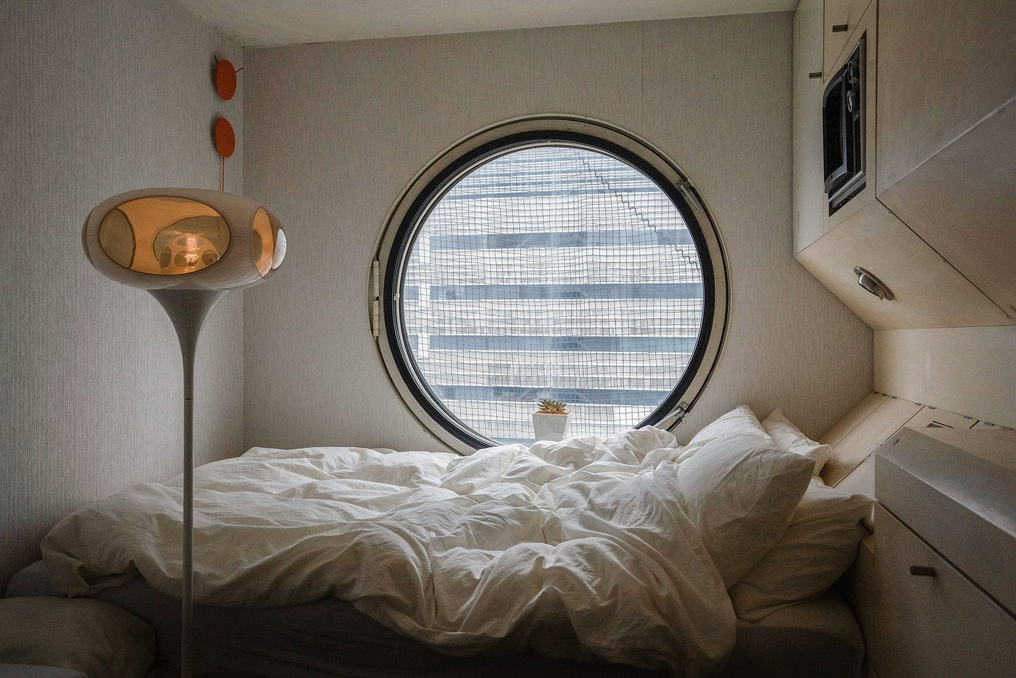 The interior of one of the Nakagin capsules, looking toward its iconic porthole window. Under the window is a bed that stretches the width of the room, with a killer Jetsons style floor lamp on one side and built-in cabinets on the other. 