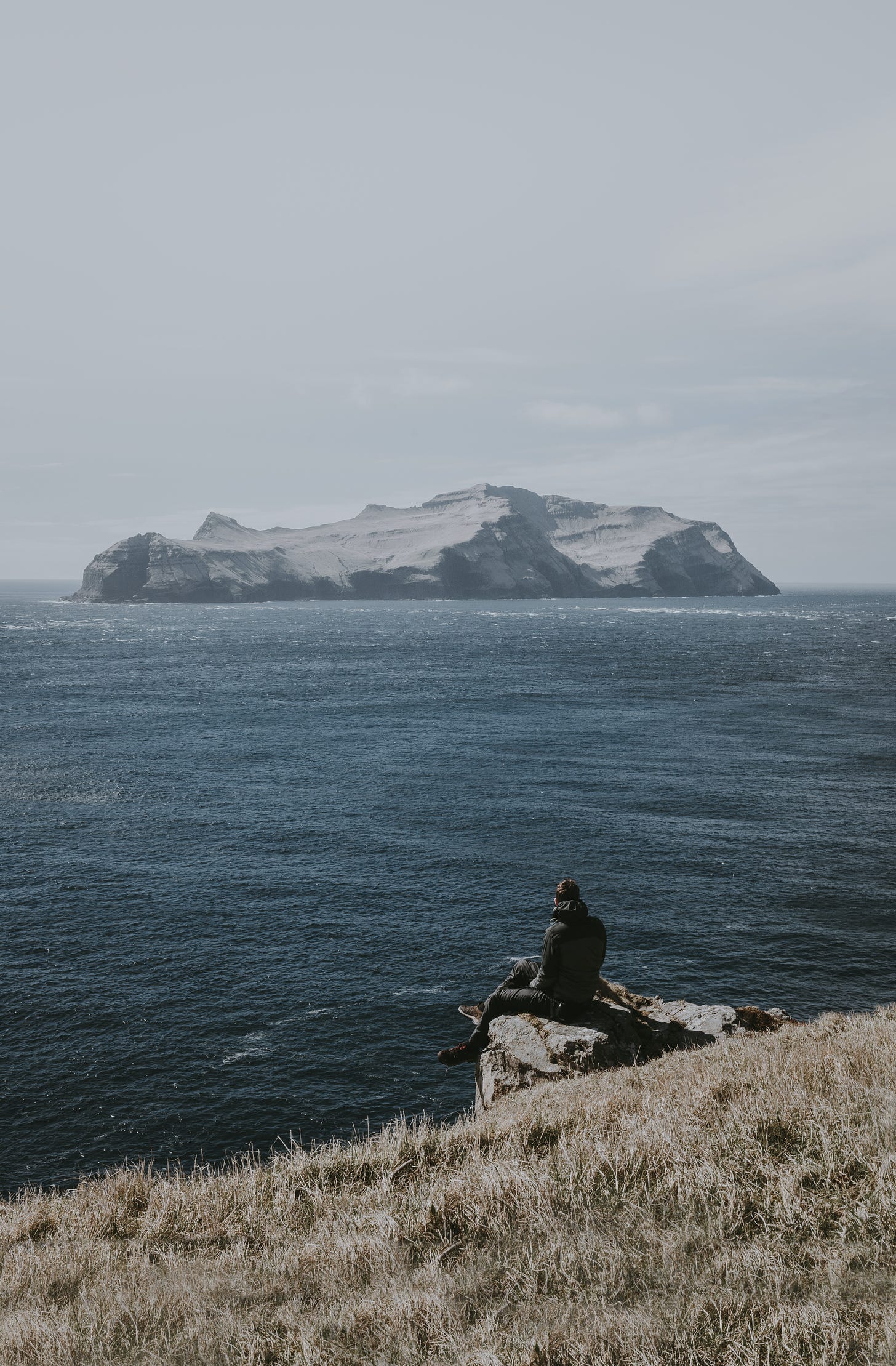 A man sitting on the edge of a cliff, looking out to a big rock in the ocean
