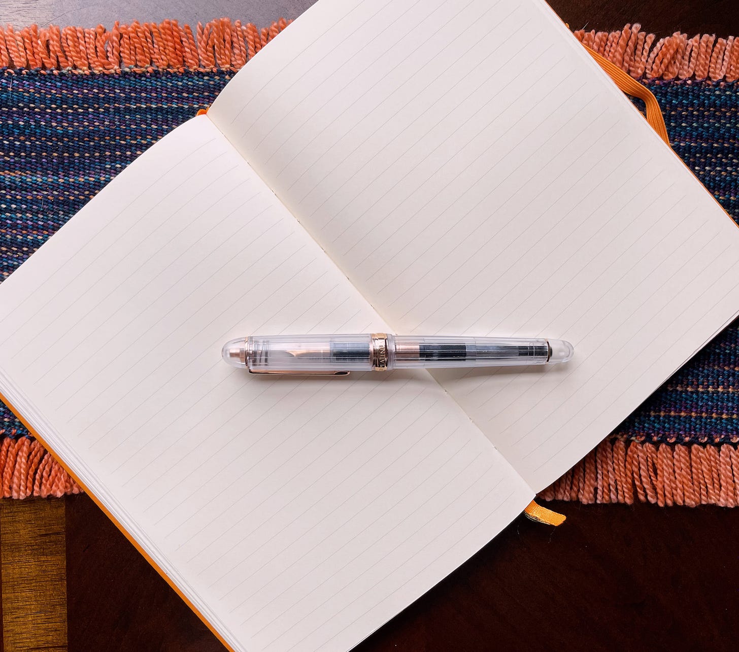 An open notebook with a pen on top, sitting on a strip of woven fabric