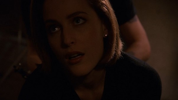 An image of Scully in a dark room, leaning forwards towards us, with an odd expression on her face.