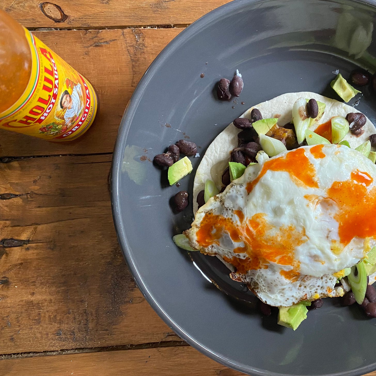 Grey bowl with toasted tortilla, black beans, avocado, eggs, a bottle of cholula hot sauce to the side
