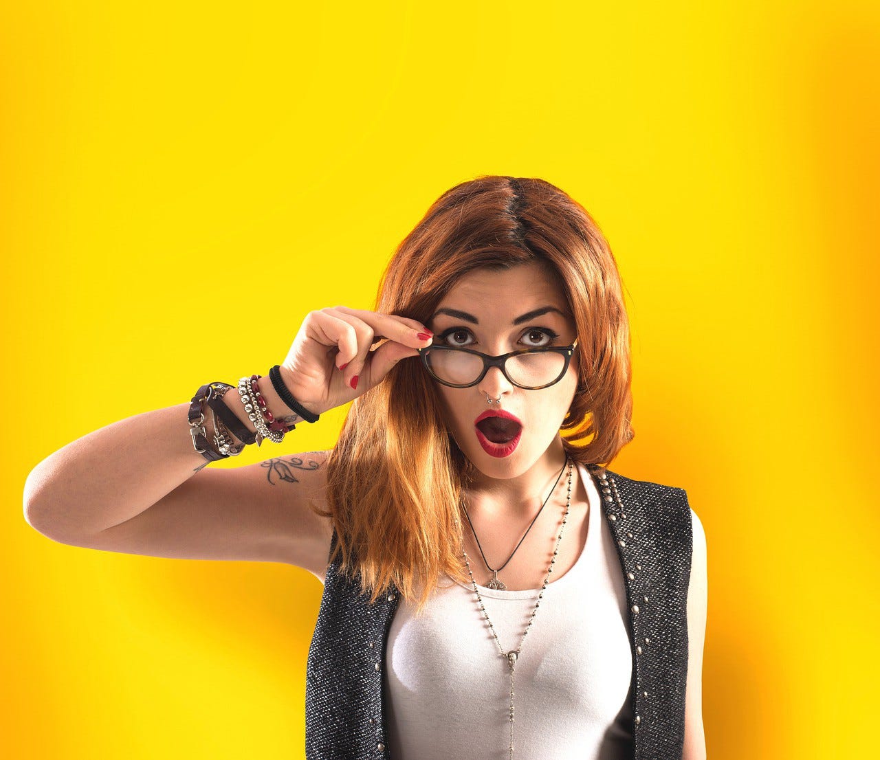 A red-haired woman wearing glasses in front of a yellow background, looking astonished