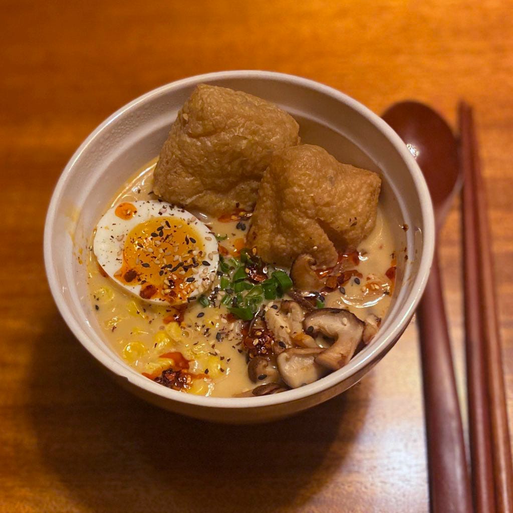 A white bowl of ramen in a creamy broth. On top of the noodles are half a boiled egg, two tofu puffs, a small pile of sliced fried mushrooms, green onions, and corn. Chili oil and sesame seeds are scattered over the top.
