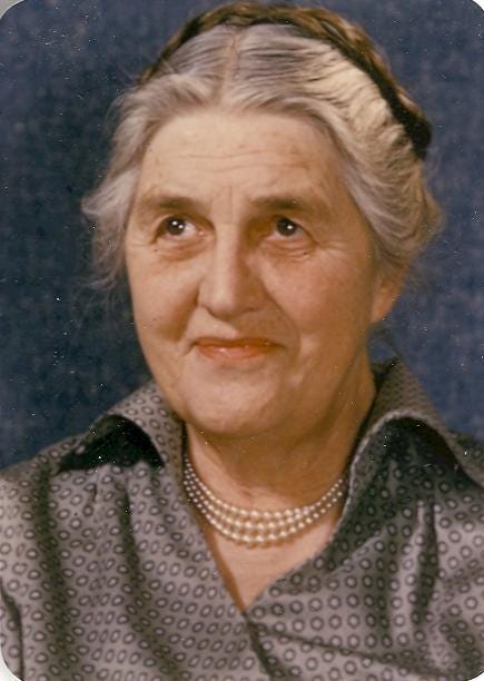 Older photo of an old woman with white skin and grey hair. She is smiling with mouth closed and looking off to the side of the camera.