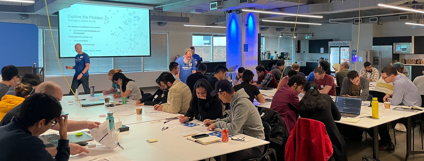 Workshop attendees at tables in pairs, working through worksheets. Projector displays, "explore the problem" with the four rules of Conway's Game of Life.