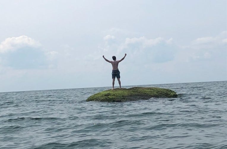 A person stands on a large rock, rising out of the waves. The rock is deep in open water. A hazy, cloudy sky dominates the upper half of the frame